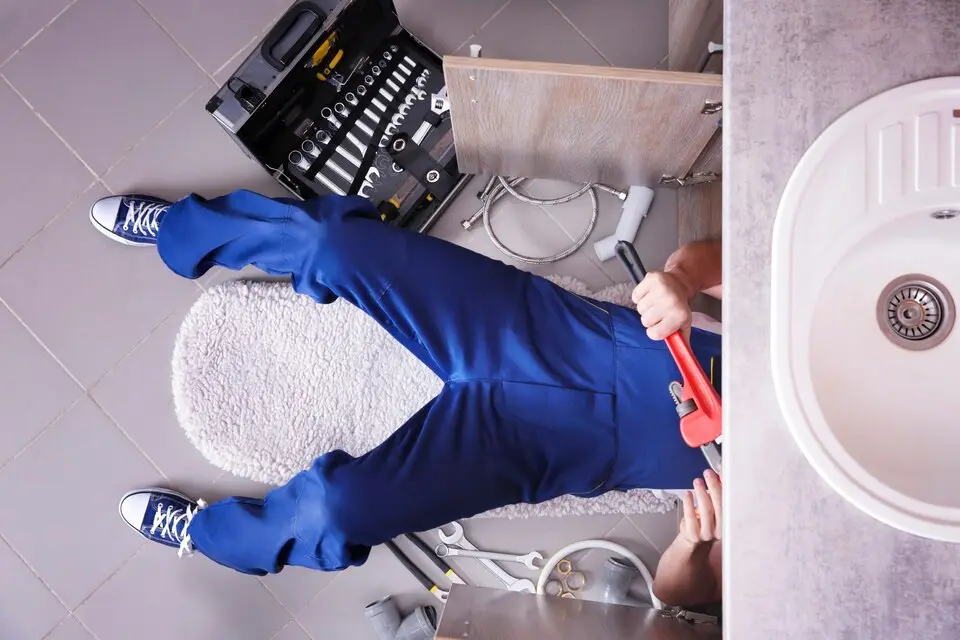 Plumber in blue overalls using a pipe wrench under a sink, surrounded by tools and plumbing components, illustrating professional plumbing maintenance and installation services.
