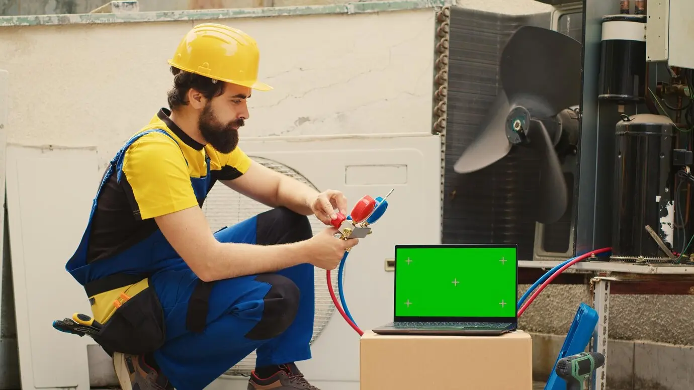 HVAC technician in yellow hard hat and blue overalls using tools to service an air conditioning unit, with a laptop displaying a green screen nearby.