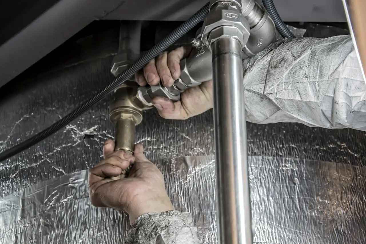 Plumber repairing water heater piping with metal fittings under a home, emphasizing plumbing services and maintenance.