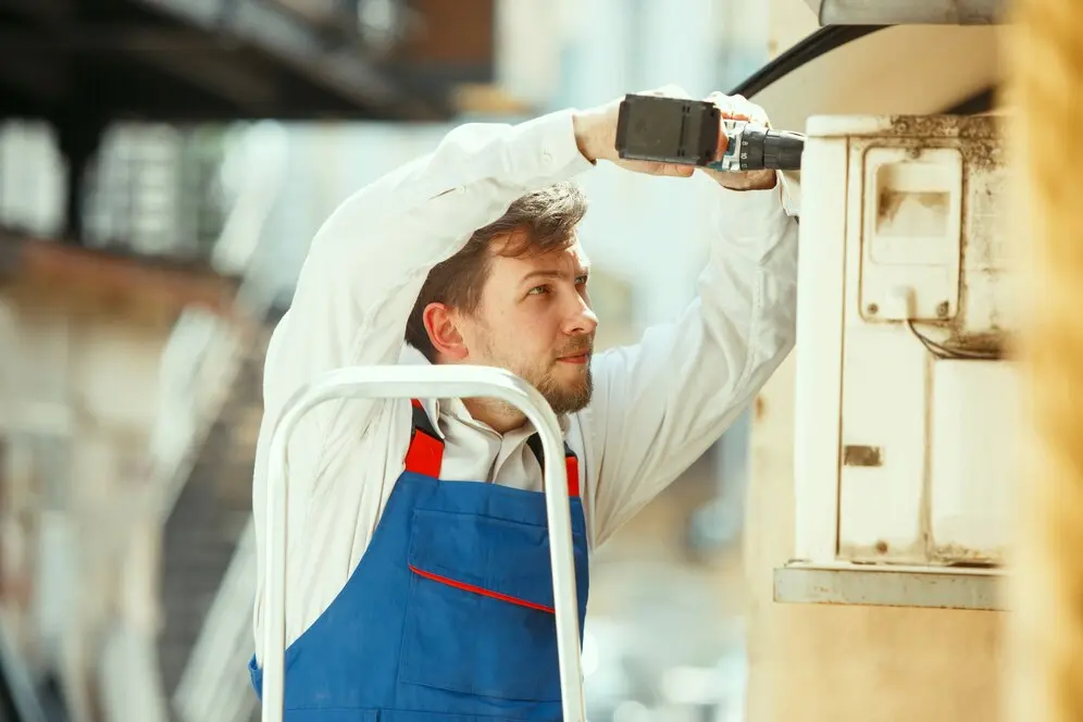 Technician performing AC maintenance using a power drill while standing on a ladder, emphasizing professional HVAC services.