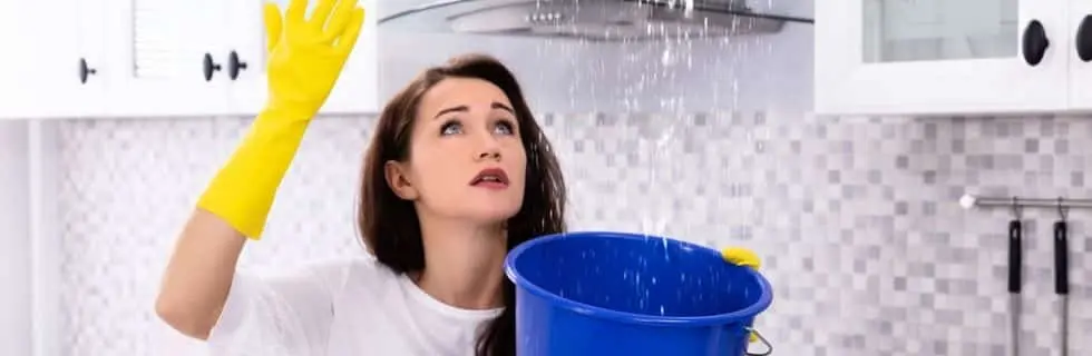 Woman in yellow gloves looking up at a water leak from a kitchen ceiling, holding a blue bucket to catch the dripping water, emphasizing the urgency of fixing leaks.