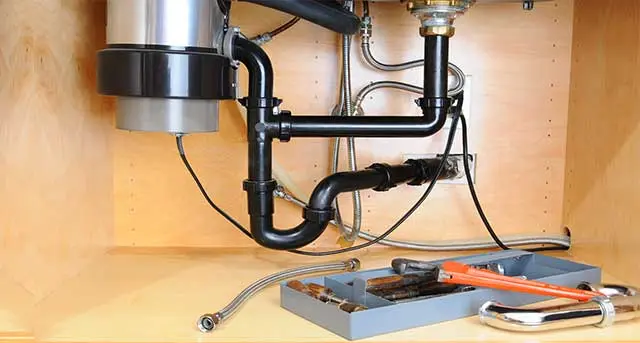 Garbage disposal unit and plumbing pipes under kitchen sink, showcasing tools for maintenance and repair services by Jim Wagner Plumbing, Inc.