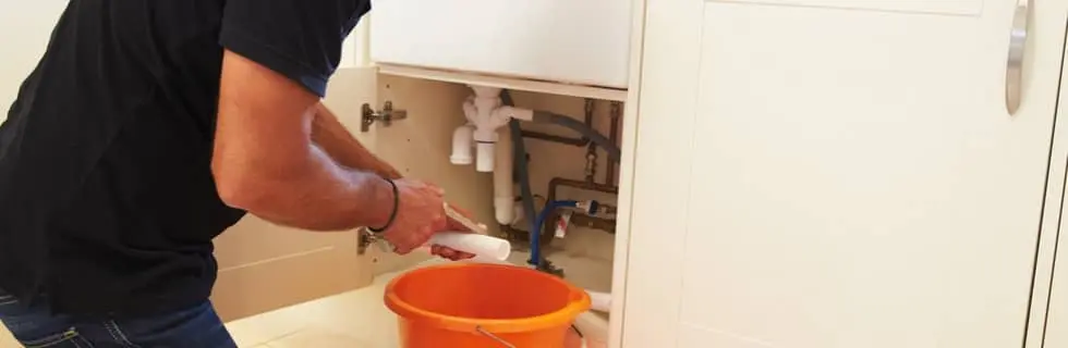 Man working under a sink with plumbing pipes, attempting a DIY repair, with an orange bucket positioned to catch water, reflecting common plumbing challenges faced by fathers.