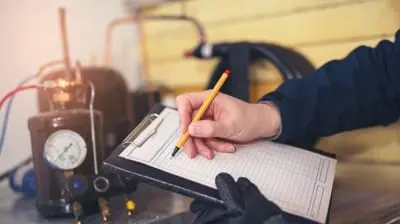 A person holding a pencil and writing on a clipboard near industrial equipment and pressure gauges, diligently performing AC maintenance in California.