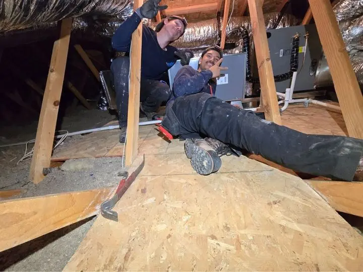 Two HVAC technicians smiling and posing in an attic, surrounded by air conditioning equipment and ductwork, illustrating AQS Heating & Air Conditioning's professional installation services.