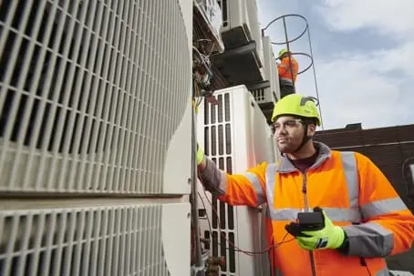 Two workers in high-visibility clothing and hard hats are performing local HVAC repair outdoors in California; one tests an HVAC unit with a meter while ensuring proper AC maintenance.
