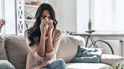 Woman sitting on a couch, holding a tissue to her nose, experiencing discomfort likely due to indoor allergens or poor air quality.