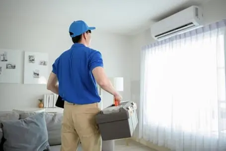 HVAC technician in blue uniform holding a toolbox, preparing to service an air conditioning unit in a bright, modern living room with large windows.