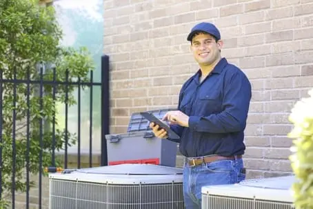 HVAC technician smiling and using a tablet next to air conditioning units, representing professional AC maintenance services by AQS Heating & Air Conditioning.