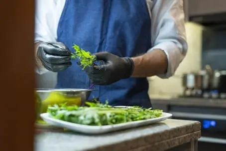 A person wearing black gloves and a blue apron prepares fresh greens in a California kitchen.