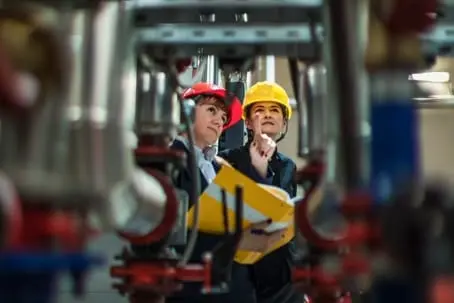 Two people in safety helmets inspect equipment in an industrial setting, one holding a clipboard and pointing. The AC technician carefully reviews the details, ensuring all aspects of AC maintenance are up to code.