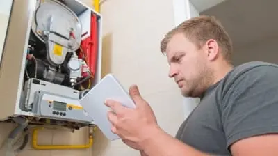 A technician from the local HVAC repair service in Hemet inspects a white gas boiler with a tablet in hand, dressed in a grey t-shirt. The boiler panel is open, revealing its inner workings.