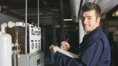 Technician in a blue uniform taking notes on a clipboard next to a panel of gauges and pipes during AC maintenance in a mechanical room.