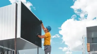 Worker in yellow shirt and blue hard hat aligns a large metal panel against a bright blue sky with white clouds, showcasing a typical day in local hvac repair work under the sunny California skies.