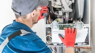 A technician in California, wearing red gloves and a cap, meticulously repairs an electrical component in an appliance during routine AC maintenance.