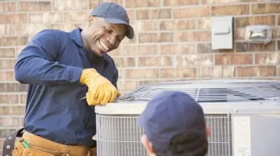 Two ac technicians service an outdoor HVAC unit. One, with a helmet on, is smiling and using a tool, while the other looks on.