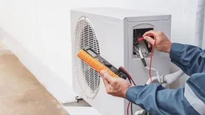 Technician performing AC maintenance in California, using a multimeter to check an outdoor air conditioning unit's electrical components.