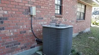 An AC technician installs an air conditioning unit outside a red brick house in California, connecting it seamlessly to an electrical box mounted on the wall.