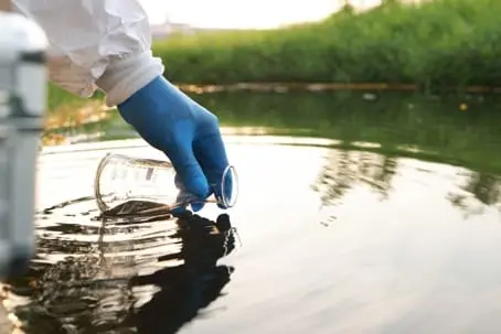 Person wearing gloves collecting water sample in a natural setting, emphasizing environmental monitoring and water quality analysis.