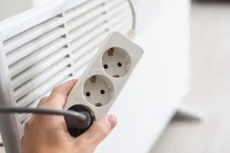 Hand holding a power strip with multiple outlets, positioned near an air conditioning unit, emphasizing the importance of proper electrical connections for HVAC maintenance.
