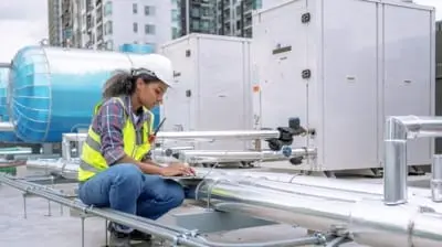 Engineer in a hard hat and safety vest working on a laptop near industrial pipes and machinery on a rooftop in California.