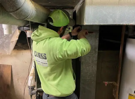 A worker in a green hoodie works on HVAC maintenance in an industrial setting with tools and machinery.