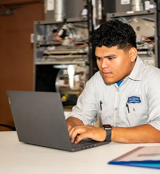 Technician in Acosta uniform working on laptop at a table, HVAC equipment in background, reflecting professional heating and cooling services.