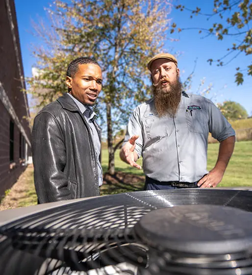 Two men discussing HVAC maintenance near an outdoor unit, one wearing a leather jacket and the other in an Acosta Heating, Cooling & Electrical uniform, surrounded by trees and blue sky.