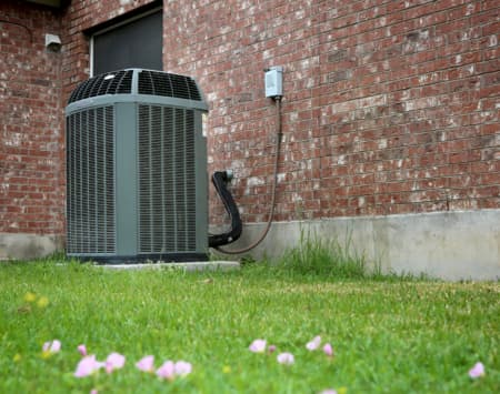 Air conditioning unit installed outside a brick home, surrounded by grass and flowers, emphasizing HVAC maintenance importance for seasonal efficiency.