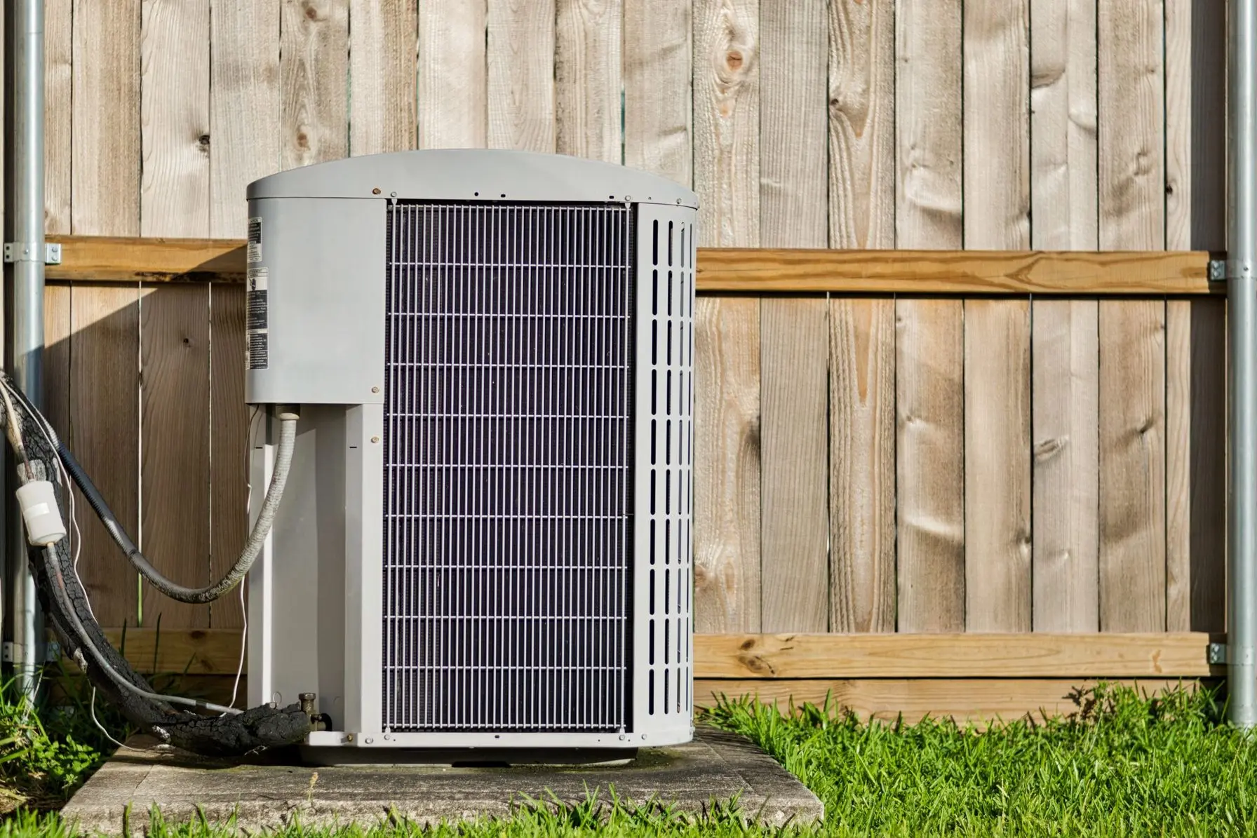 Central air conditioning unit in a residential backyard in front of a defocused wooden fence and lawn area