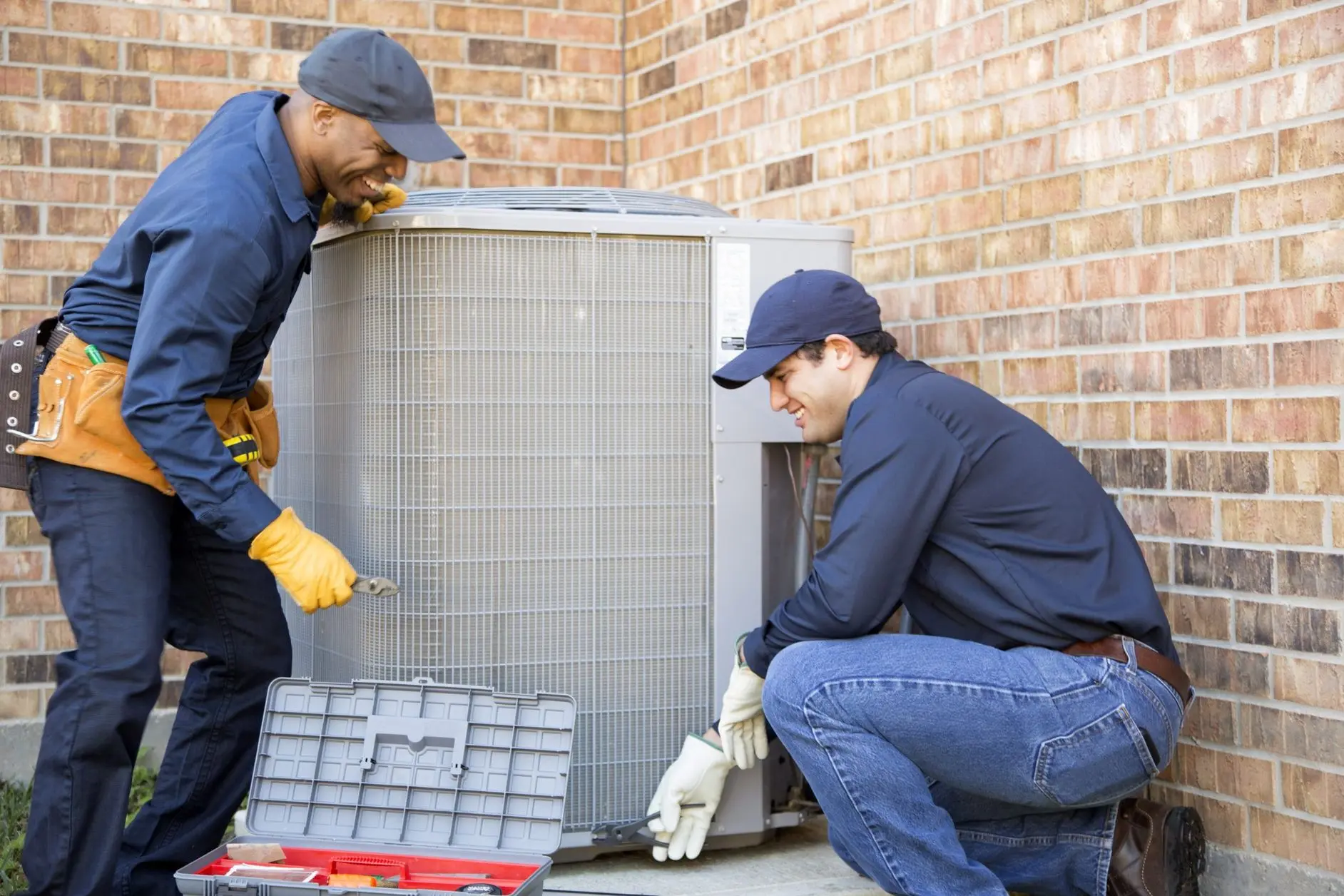 Two HVAC technicians performing maintenance on an outdoor air conditioning unit, equipped with tools and wearing gloves, showcasing the importance of regular system upkeep for optimal performance.