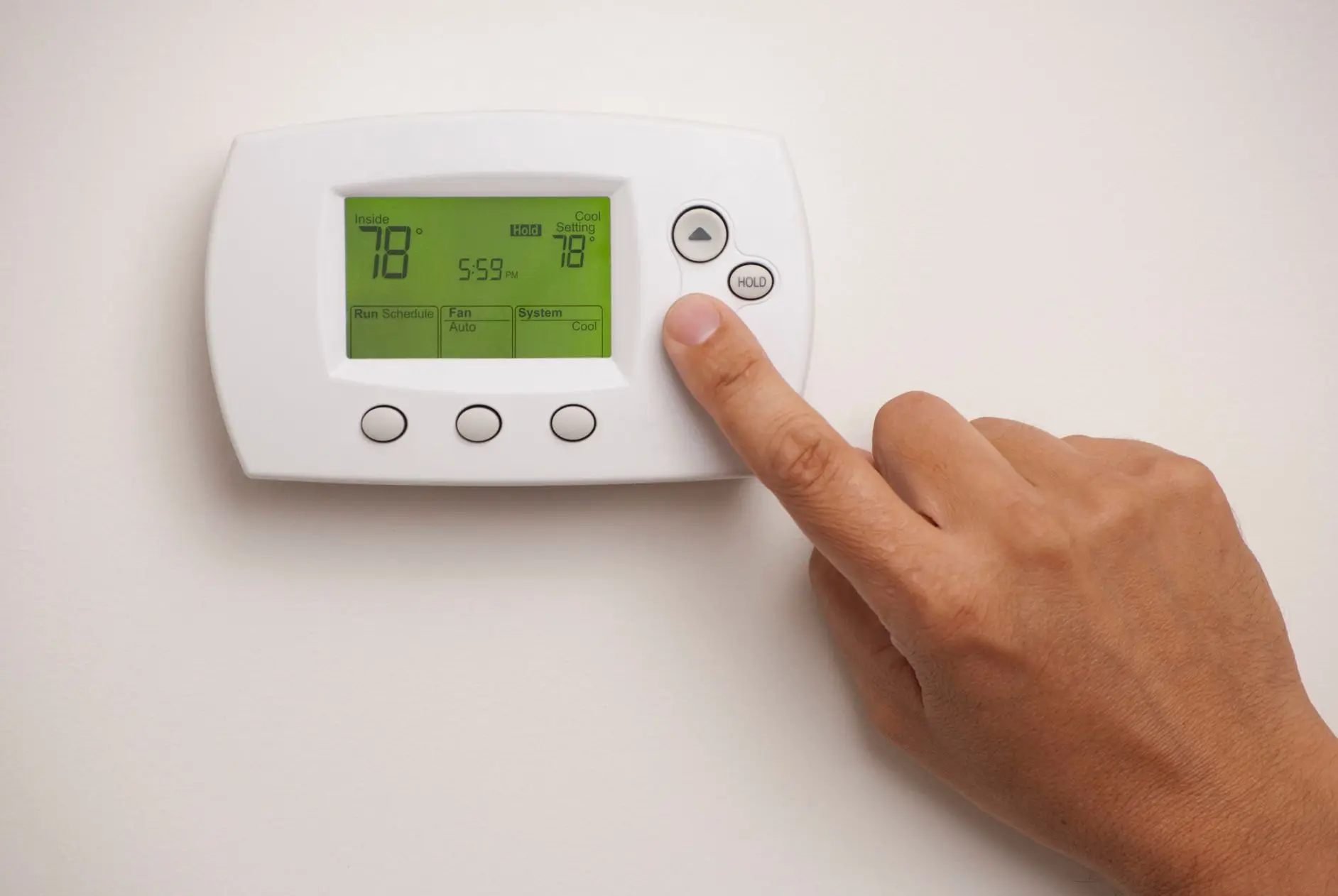 Hand adjusting a digital thermostat displaying indoor and cooling temperatures, relevant to HVAC system settings and energy efficiency discussions.