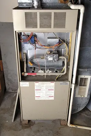 Old furnace in a basement showing exposed wiring, control panel with safety warnings, and ventilation pipes for HVAC system.