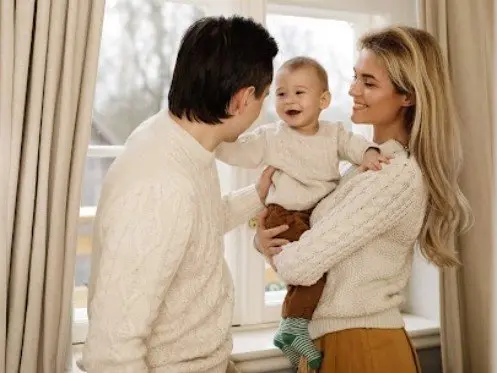 Family enjoying time together indoors, smiling with a baby, wearing cozy sweaters near a window, highlighting warmth and comfort in a home setting.