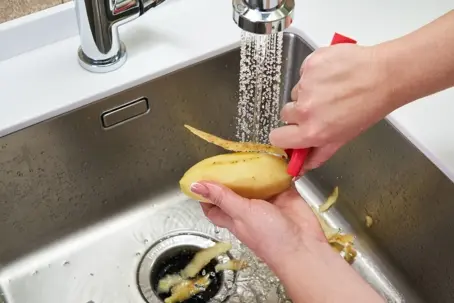 Close-up of hands peeling a raw potato with a red peeler over a modern sink with running tap water.