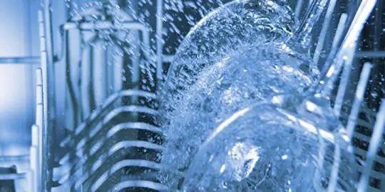 Inside view of an operating dishwasher with powerful water jets spraying over clear glass plates and a sliding dish rack.
