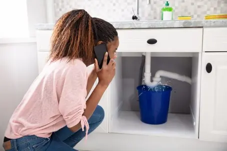 A stressed homeowner calling a plumber or emergency repair service to fix a sudden plumbing leak under the kitchen counter.