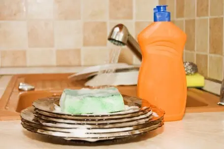 A soapy green sponge sitting on top of a stack of dirty dishes next to an orange bottle of dish soap by a kitchen sink.