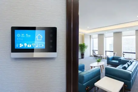 A wall-mounted touchscreen smart thermostat and home security monitor panel with a modern office lounge visible in the background.