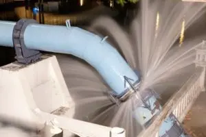 Massive water leak bursting from a ruptured blue industrial pipeline connection next to a concrete support structure.