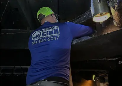 HVAC technician from License to Chill performing maintenance work in a dimly lit space, wearing a blue branded shirt and a green cap.