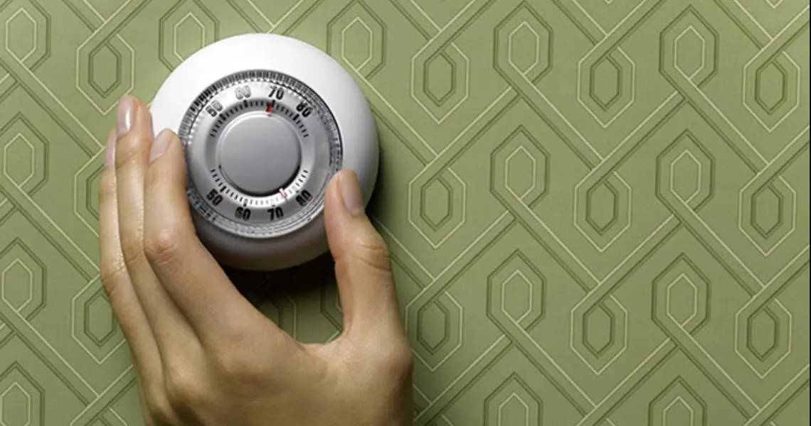 Hand adjusting a thermostat on a patterned green wall, emphasizing home heating efficiency and winter preparation.