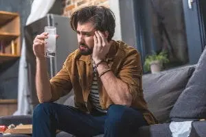 Man sitting on a couch holding a glass of water, appearing distressed, illustrating the effects of hard water on health and comfort in home plumbing.