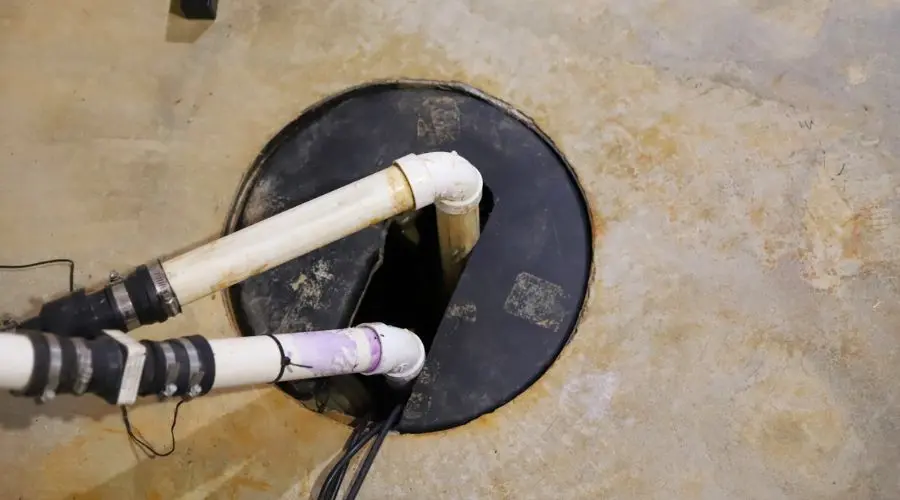 Two white plumbing pipes leading to a circular hole in the concrete, with a black cover showing signs of wear.