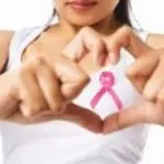 A woman forming a heart shape with hands, wearing a white tank top with a pink ribbon for breast cancer awareness.