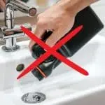 Person pouring liquid from a black bottle into a kitchen sink with faucet and drain, marked by a red prohibition "X".