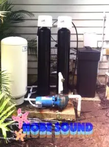 Water softening system installation featuring two black water softeners, a white pressure tank, and a blue pump, surrounded by greenery, relevant to Allore's Plumbing Services in Stuart.