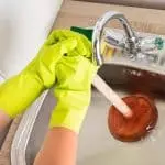 Bright yellow rubber gloves cleaning a kitchen sink with a wooden plunger for effective household maintenance.