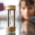 Wooden hourglass showcasing flowing sand, with a blurred figure representing contemplation in the plumbing services context.