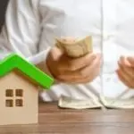 Person counting banknotes by a model house with a green roof, depicting financial planning for home maintenance.